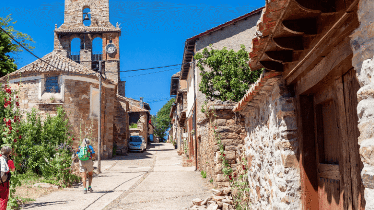 Pellegrini lungo una strada di pietra nel villaggio di Rabanal del Camino, sul Cammino di Santiago, con chiesa romanica e case in pietra sotto il cielo azzurro.