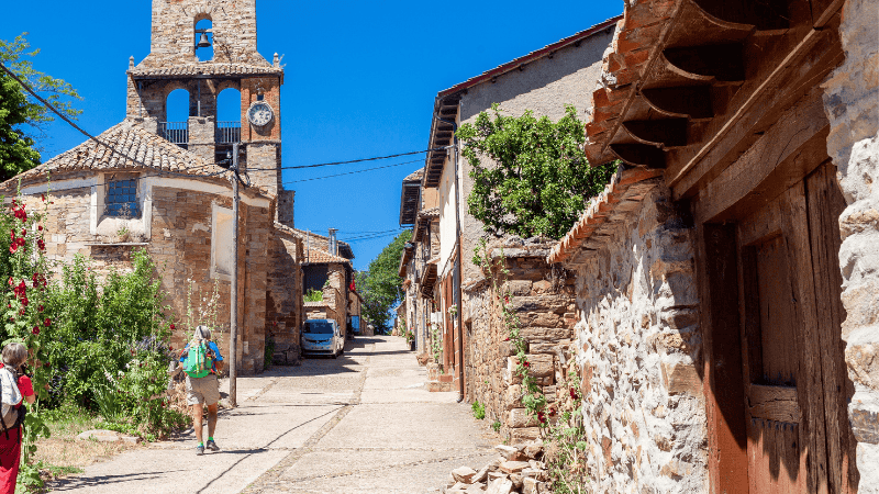 Pellegrini lungo una strada di pietra nel villaggio di Rabanal del Camino, sul Cammino di Santiago, con chiesa romanica e case in pietra sotto il cielo azzurro.