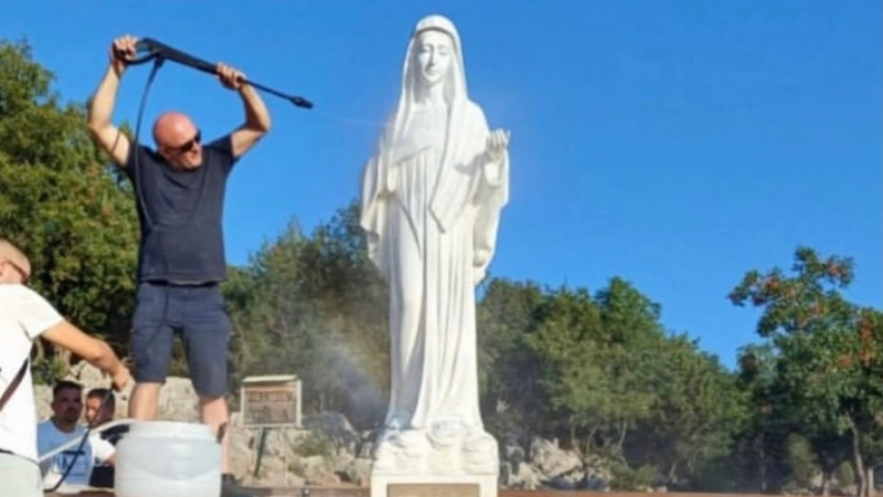 Un uomo lava con l'idropulitrice la statua bianca della Madonna di Medjugorje contro il cielo azzurro